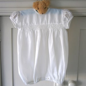 May include: A white baby romper with short, puffed sleeves and a smocked bodice. The romper is hanging on a hanger with a teddy bear head. The fabric is gathered at the waist and ankles, creating a bubble shape.