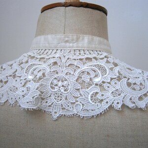 Puede incluir: Un cuello de encaje blanco con un intrincado dise&ntilde;o floral se muestra en un maniqu&iacute;. El cuello est&aacute; hecho de encaje delicado y calado y tiene un borde festoneado. El cuello est&aacute; sujeto a una banda de tela blanca.