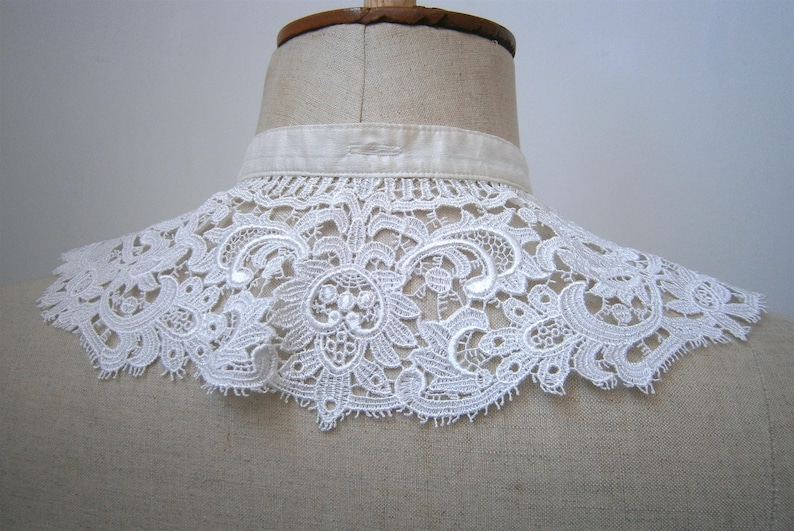 Puede incluir: Un cuello de encaje blanco con un intrincado dise&ntilde;o floral se muestra en un maniqu&iacute;. El cuello est&aacute; hecho de encaje delicado y calado y tiene un borde festoneado. El cuello est&aacute; sujeto a una banda de tela blanca.