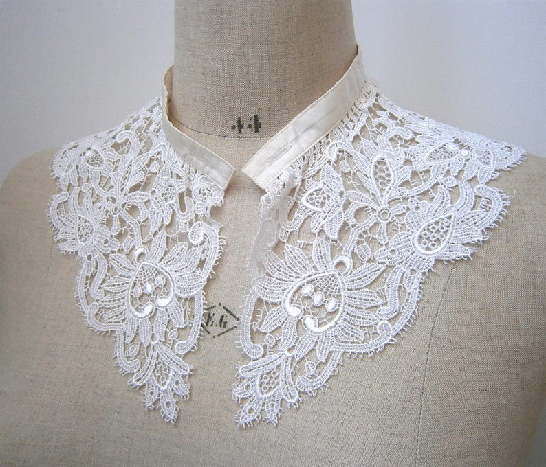 Puede incluir: Un cuello de encaje blanco con un intrincado dise&ntilde;o floral. El cuello est&aacute; hecho de encaje delicado y presenta un escote de cinta de sat&eacute;n. El cuello se exhibe en un maniqu&iacute;, mostrando su estilo elegante y vintage.