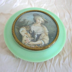 May include: A round, light green trinket box with a gold-rimmed, circular lid. The lid features a miniature painting of a woman in a hat holding flowers. The box has a smooth, glossy finish, and the painting has soft, muted colors.