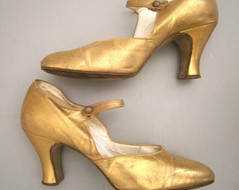 May include: A pair of metallic gold Mary Jane heels featuring a rounded toe, a single strap across the instep with a small circular button closure, and a curved mid-height heel. The interior lining is white, and the exterior has a smooth finish.