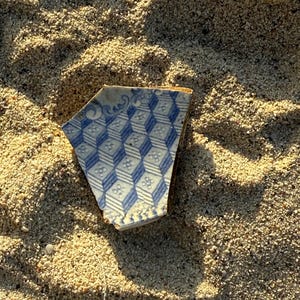 Könnte beinhalten: Ein blaues und weißes Keramikplattenfragment mit geometrischem Muster, teilweise im Sand vergraben.