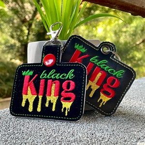 以下が含まれることがあります： 緑、黄色、赤の滴るデザインで「black king」と書かれた、黒い革製のキーホルダー2個
