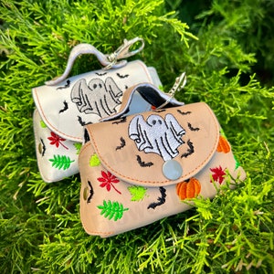 May include: Two small, fabric coin purses with zipper closures. Both purses feature an embroidered ghost design and a fall-themed pattern with leaves, pumpkins, and bats. The purse on the left is white with a silver zipper pull, and the purse on the right is beige with a gold zipper pull.