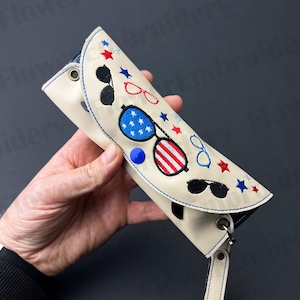 Puede incluir: Un estuche para gafas de cuero blanco con un diseño de bandera estadounidense azul, rojo y blanco y varios pares de gafas de sol bordados. El estuche tiene una correa azul con un cierre plateado.