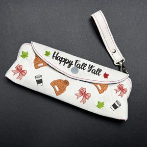 May include: White eyeglasses case with a wrist strap. The case is decorated with embroidered fall-themed images, including a beanie, bow, coffee cup, and maple leaf. The text "Happy fall y'all" is embroidered on the front.