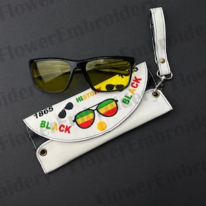 May include: Black sunglasses with yellow lenses rest on a white glasses case. The case features embroidered text and graphics, including the words "BLACK" and "HISTORY 1865". A white wrist strap is attached to the case.