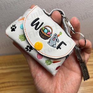 May include: White leather key chain with a bunny, a basket of Easter eggs, and the word "Woof" embroidered on it. The key chain has a yellow button and a silver key ring.