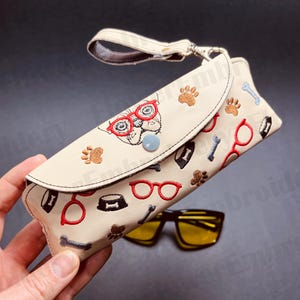 May include: A beige eyeglasses case with a zip closure and a strap. The case is decorated with an embroidered design of a dog wearing glasses, bones, and paw prints. The case is open and a pair of yellow sunglasses are visible in the background.
