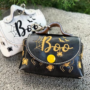 Könnte beinhalten: Zwei schwarze und weiße Halloween-Taschen mit Reißverschluss und einem "Boo"-Stickmotiv. Die Taschen haben ein Spinnennetz- und Fledermausdesign. Eine Tasche ist weiß mit schwarzer Stickerei und die andere ist schwarz mit goldener Stickerei.