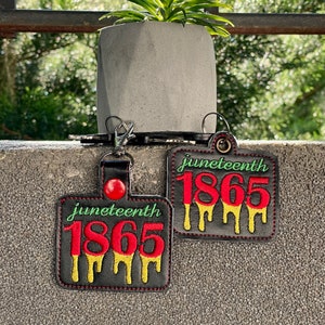 May include: Two black leather keychains with red stitching. Each keychain features a red and yellow dripping number "1865" with the word "Juneteenth" above it.