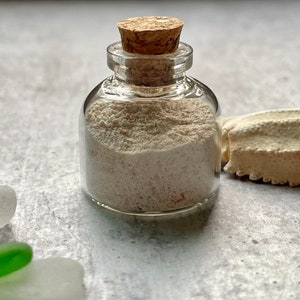 May include: A small, clear glass bottle filled with light-colored sand, sealed with a cork stopper. The bottle sits on a gray surface, with pieces of sea glass and a seashell nearby. The sand appears to have a slightly pink hue.
