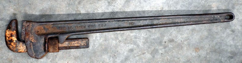 May include: A vintage, rusty pipe wrench with a long handle. The wrench is dark gray with areas of rust and the text "THE RIDGE TOOL CO. ELYRIA OHIO U.S.A." is visible. The adjustable jaw is open, ready for use. A classic tool for plumbing or mechanical work.