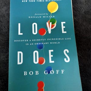May include: A teal-covered book titled "LOVE DOES" by Bob Goff. The cover features the title in large white letters, with colourful balloons. The text "NEW YORK TIMES BESTSELLER" is at the top. The book's subtitle is "DISCOVER A SECRETLY INCREDIBLE LIFE IN AN ORDINARY WORLD".