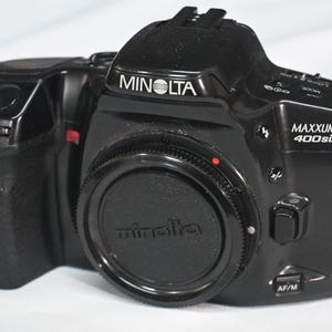 Puede incluir: Una cámara de película de 35 mm Minolta Maxxum 400si negra con una tapa de lente negra. La cámara tiene un logotipo Minolta plateado en la parte superior y un logotipo Maxxum 400si plateado en el lateral.