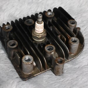 May include: A close-up of a silver engine cylinder head with a Champion spark plug inserted. The cylinder head has multiple holes for spark plugs and is designed for a multi-cylinder engine.