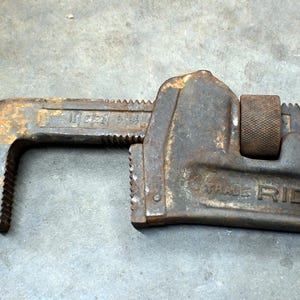 May include: Close-up of a rusty, vintage RIDGID pipe wrench. The wrench is made of dark metal with visible wear and tear, including rust. The text "TRADE RIDGID" is visible on the side.