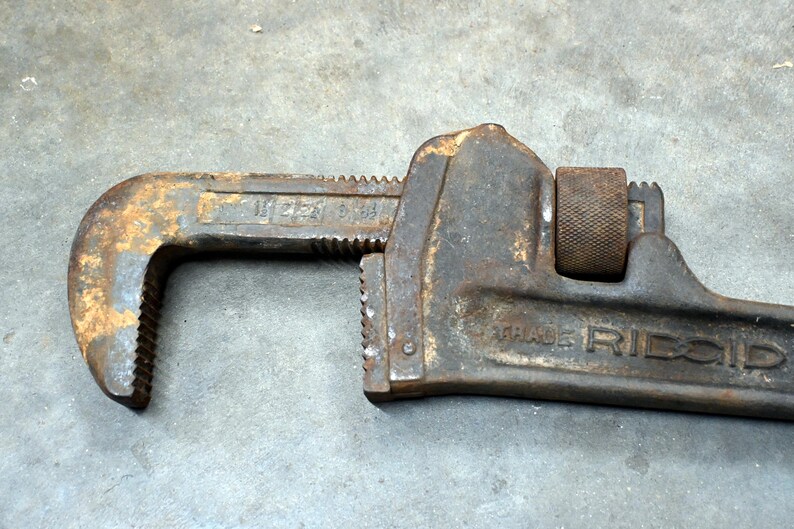 May include: Close-up of a rusty, vintage RIDGID pipe wrench. The wrench is made of dark metal with visible wear and tear, including rust. The text "TRADE RIDGID" is visible on the side.
