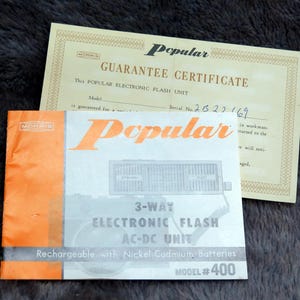 May include: A vintage "Popular" electronic flash unit with a guarantee certificate. The flash unit is a 3-way AC-DC model, rechargeable with nickel cadmium batteries. The model number is #400. The guarantee certificate is from Morris.