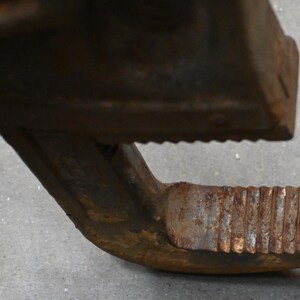 May include: Close-up of a rusty, heavy-duty metal clamp. The clamp has a textured, ridged jaw and a curved handle with similar ridges. The metal shows signs of wear and age, with a weathered, brown appearance.