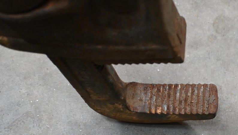 May include: Close-up of a rusty, heavy-duty metal clamp. The clamp has a textured, ridged jaw and a curved handle with similar ridges. The metal shows signs of wear and age, with a weathered, brown appearance.