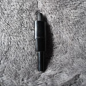 May include: A black, cylindrical bottle stopper with a ridged top. The stopper is made of a glossy material and has a simple, elegant design. The background is a soft, gray, textured fabric.