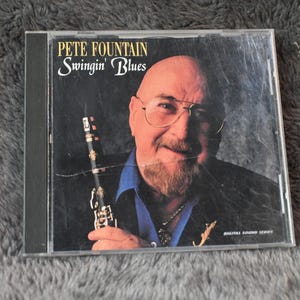Puede incluir: Caja de CD con Pete Fountain, con el título "Swingin' Blues". La portada muestra un primer plano de Fountain, con gafas y sosteniendo un clarinete. El fondo es oscuro y el texto es dorado.