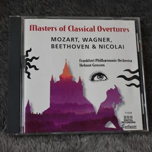 May include: A CD case featuring "Masters of Classical Overtures" with the names Mozart, Wagner, Beethoven & Nicolai. The cover art includes a purple silhouette of a building and an eye. The CD is a digital recording.