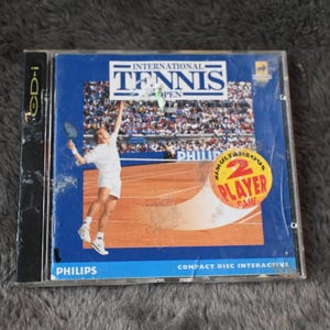 May include: A CD-i game case for "International Tennis Open" featuring a tennis player in action. The cover is blue with the game title in white and a yellow circle indicating a 2-player game. The Philips logo is visible.