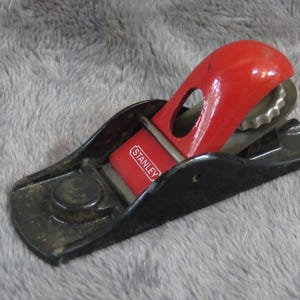 May include: A red and black Stanley block plane. The plane has a red handle and a black base. The word "STANLEY" is printed on the red lever cap. This tool is used for smoothing and shaping wood.