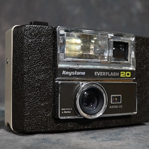 May include: A black and silver Keystone Everflash 20 camera with a built-in flash. The camera has a 40mm f/5.6 lens and an electric eye.