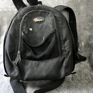 May include: Black backpack with a front zippered pocket, mesh pocket, and adjustable straps. The backpack has the "lowepro" logo and a small bear emblem. The bag is made of a durable, textured material.