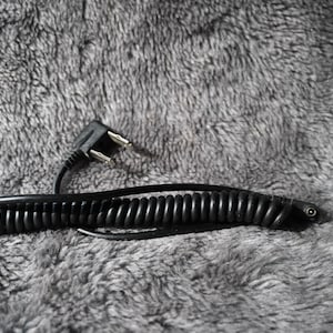 May include: A black coiled cord with a two-prong plug. The cord is lying on a gray, textured surface. The cord is a spiral shape and appears to be made of rubber or plastic.