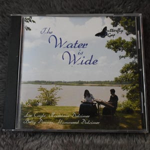 Puede incluir: Caja de CD con el título del álbum "The Water is Wide" en escritura azul. La portada presenta una vista panorámica de un lago con dos personas tocando instrumentos. También se ve el texto "Butterfly Gap" y los nombres de los artistas.