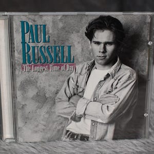 Può includere: Una custodia per CD con una foto in bianco e nero di un uomo con capelli castano corti. Il testo sulla custodia recita "Paul Russell The Longest Time of Day".