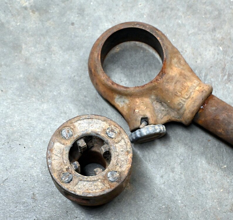 以下が含まれることがあります： 錆びたパイプねじ切り工具セット。ダイヘッドとハンドルが含まれています。ダイヘッドは円形で、複数の切削スロットがあり、ハンドルには大きなアイレットが付いています。工具は金属製で、使用感が見られます。