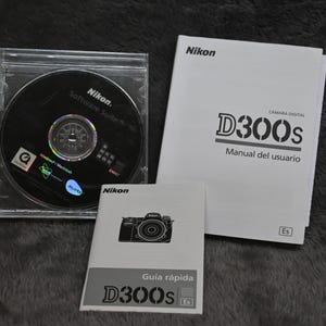 Könnte beinhalten: Ein Nikon D300s Kamera-Kit, bestehend aus einer Software-CD in einer transparenten Hülle, einem Benutzerhandbuch und einer Kurzanleitung. Die Handbücher sind weiß mit schwarzem Text, die CD ist schwarz mit silbernen und blauen Akzenten.