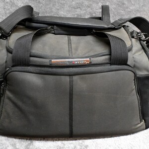 May include: A dark gray travel bag with black accents. The bag has a top handle, a shoulder strap, and a front zippered pocket. The bag has the word "DELSEY" on the front. The bag is made of a durable material.