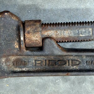 May include: A well-worn, rusty pipe wrench with the brand name "RIDGID" visible. The wrench is primarily dark gray with areas of rust and a blue handle. The adjustable jaw and toothed section are prominent.