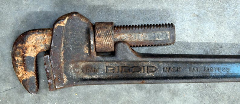 May include: A well-worn, rusty pipe wrench with the brand name "RIDGID" visible. The wrench is primarily dark gray with areas of rust and a blue handle. The adjustable jaw and toothed section are prominent.
