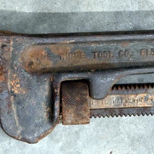 May include: An antique, rusty pipe wrench with the text "THE RIDGE TOOL CO. ELYRIA, OHIO U.S.A." stamped on the handle. The wrench is dark gray with areas of rust, showing signs of age and use. The adjustable jaws are open.