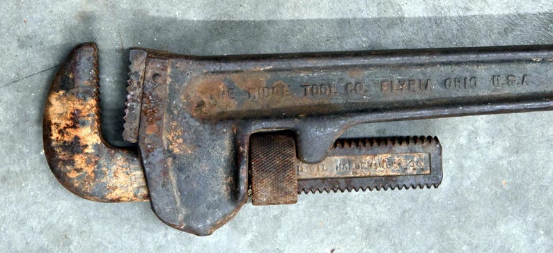 May include: An antique, rusty pipe wrench with the text "THE RIDGE TOOL CO. ELYRIA, OHIO U.S.A." stamped on the handle. The wrench is dark gray with areas of rust, showing signs of age and use. The adjustable jaws are open.