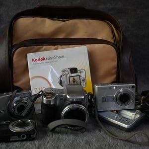 May include: A collection of digital cameras, accessories, and a carrying bag. The assortment includes several silver and black cameras, a charger, and a Kodak EasyShare manual. The bag is tan and brown.