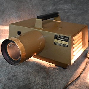 Könnte beinhalten: Vintage Filmprojektor in Goldtönen mit schwarzem Griff und Objektiv. Der Projektor hat ein zylindrisches Objektivgehäuse und einen rechteckigen Körper mit Lüftungsschlitzen. Das Etikett lautet "Seerite".