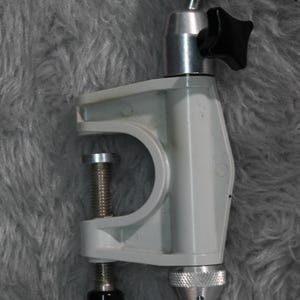 May include: A white plastic clamp with a silver metal swivel head and black knobs. The clamp has a screw to tighten it and a threaded hole for attaching a tripod or other device.