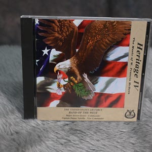 May include: CD case with a bald eagle holding a branch of green leaves against a red, white, and blue American flag background. The text on the case reads "Heritage IV The Music of W. Francis Mclleth THE UNITED STATES AIR FORCE BAND OF THE WEST Major Steven Grimes - Commander Captain Dianne Varela - Vice Commander"