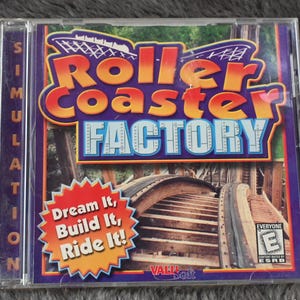 May include: A RollerCoaster Factory PC game in a clear plastic case. The cover features the game's title in orange and blue, with a roller coaster image. The text "Dream It, Build It, Ride It!" is also visible.