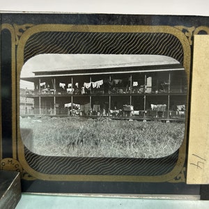 Puede incluir: Fotografía en blanco y negro de un edificio de dos pisos con porche, ropa tendida y hierba alta en primer plano. La imagen está enmarcada con un borde negro con detalles dorados. Una etiqueta con el número 14 está a la derecha.