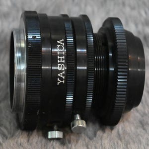 May include: A black Yashica camera lens adapter with ridged edges and a silver screw. The adapter has the brand name "YASHICA" printed in white. The adapter is designed to connect a camera lens to a camera body.